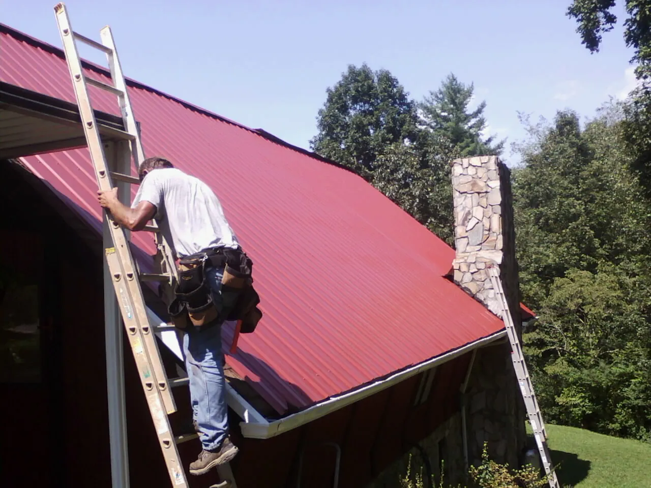 Roofer on a ladder beside a red metal roof and stone chimney — Soffit & Fascia Repair by Roofing Brothers in Nebraska City